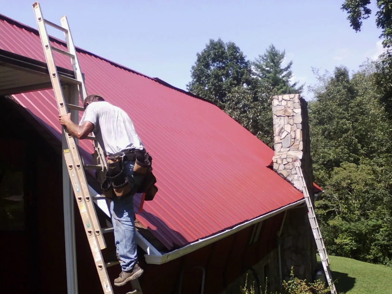 Roofer on a ladder beside a red metal roof and stone chimney — Soffit & Fascia Repair by Roofing Brothers in Herald